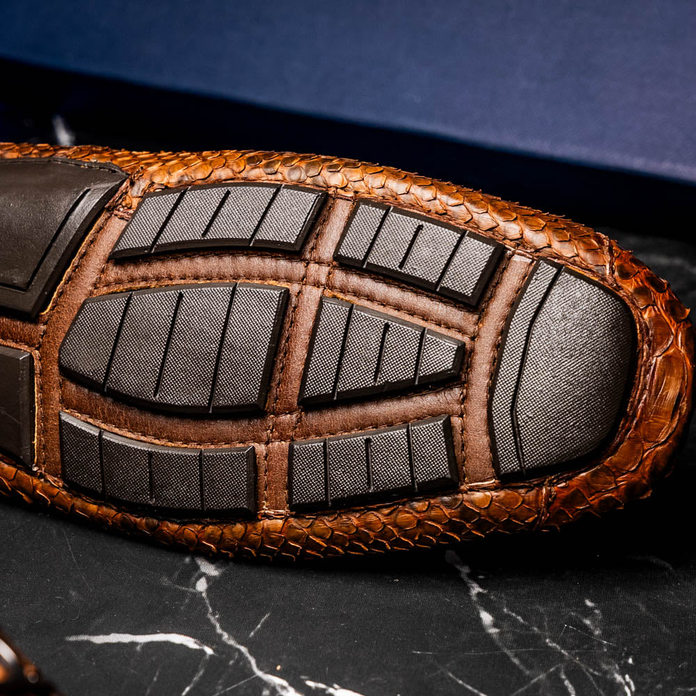 The Marco Di Milano Pia Cognac features brown python skin driver loafers with a metal buckle, visible black rubber soles, and one shoe upright while the other lies flat—a luxury men's shoe designed for style seekers.