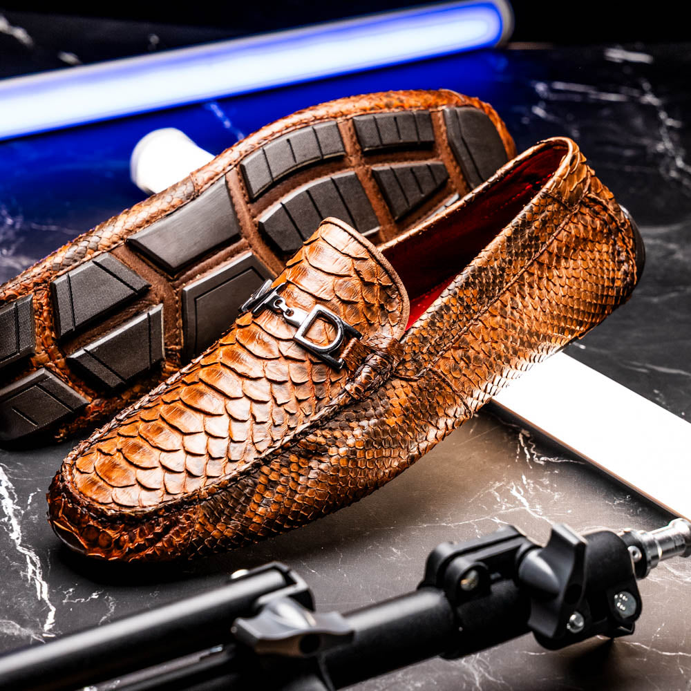 The Marco Di Milano Pia Cognac features brown python skin driver loafers with a metal buckle, visible black rubber soles, and one shoe upright while the other lies flat—a luxury men's shoe designed for style seekers.