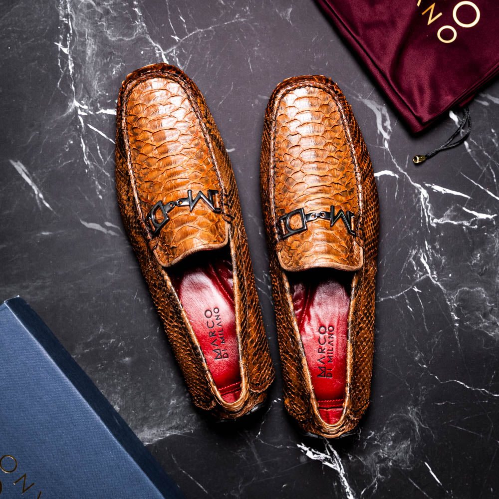 The Marco Di Milano Pia Cognac features brown python skin driver loafers with a metal buckle, visible black rubber soles, and one shoe upright while the other lies flat—a luxury men's shoe designed for style seekers.