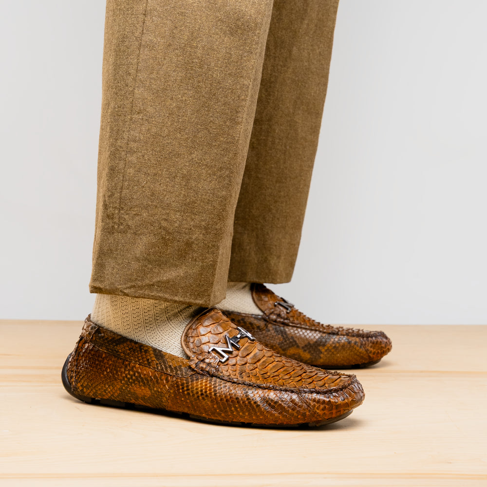 The Marco Di Milano Pia Cognac features brown python skin driver loafers with a metal buckle, visible black rubber soles, and one shoe upright while the other lies flat—a luxury men's shoe designed for style seekers.