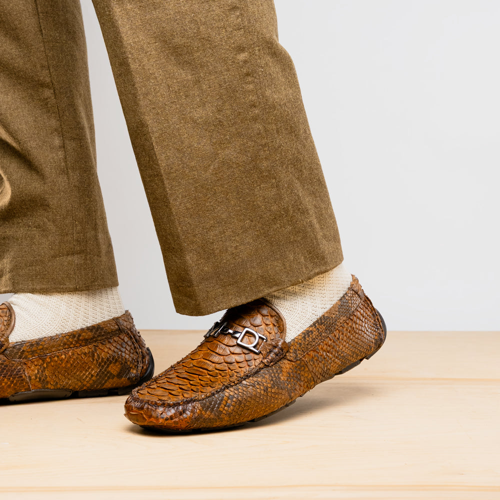 The Marco Di Milano Pia Cognac features brown python skin driver loafers with a metal buckle, visible black rubber soles, and one shoe upright while the other lies flat—a luxury men's shoe designed for style seekers.