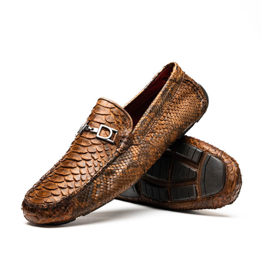 The Marco Di Milano Pia Cognac features brown python skin driver loafers with a metal buckle, visible black rubber soles, and one shoe upright while the other lies flat—a luxury men's shoe designed for style seekers.