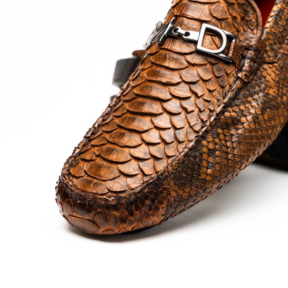 The Marco Di Milano Pia Cognac features brown python skin driver loafers with a metal buckle, visible black rubber soles, and one shoe upright while the other lies flat—a luxury men's shoe designed for style seekers.