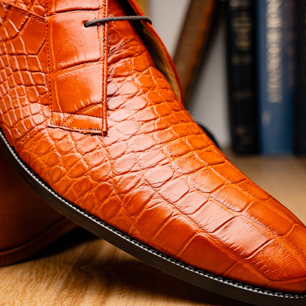 The Pietro Honey boots by Marco Di Milano are orange-brown genuine alligator, hand-painted with crocodile texture—displayed upright and sideway to show the sole—ideal for luxury footwear enthusiasts.