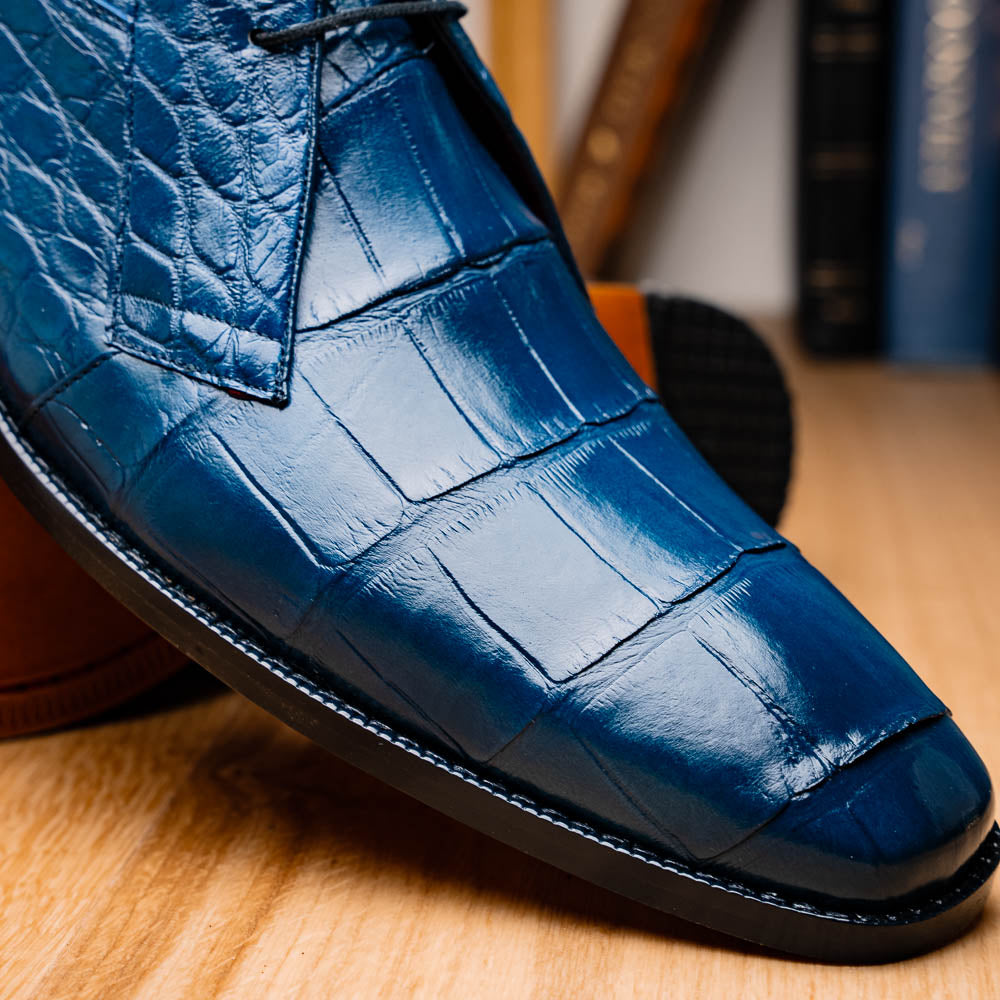 The Marco Di Milano "Pietro Blue Jeans" are luxury men's lace-up boots in blue crocodile-pattern leather, with one standing upright and the other tilted to reveal the tan embossed sole.