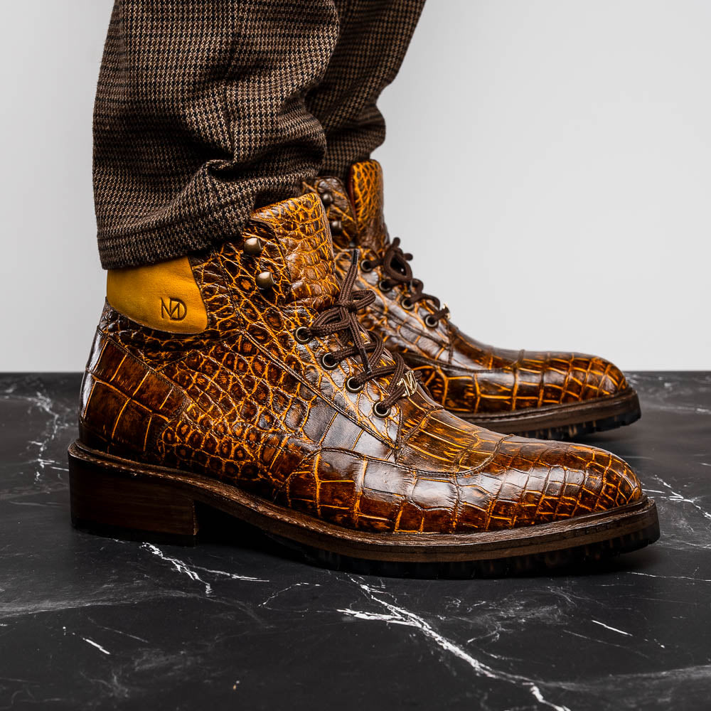 The Potenza Mustard/Brown boots by Marco Di Milano feature brown crocodile-pattern leather, lace-up fronts, tan ankle detail, and textured rubber soles with hand-painted finishes—one boot upright, the other on its side.