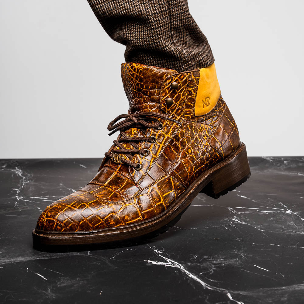 The Potenza Mustard/Brown boots by Marco Di Milano feature brown crocodile-pattern leather, lace-up fronts, tan ankle detail, and textured rubber soles with hand-painted finishes—one boot upright, the other on its side.