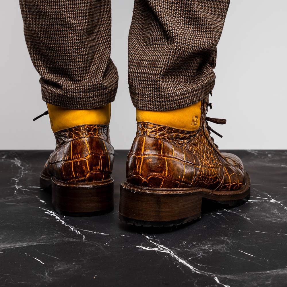 The Potenza Mustard/Brown boots by Marco Di Milano feature brown crocodile-pattern leather, lace-up fronts, tan ankle detail, and textured rubber soles with hand-painted finishes—one boot upright, the other on its side.
