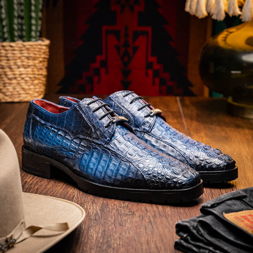 The Marco Di Milano Ragusa Stained Navy is a pair of luxury men's dress shoes in blue crocodile-patterned leather—one upright, one angled to display the sole—handcrafted for sophistication and timeless style.
