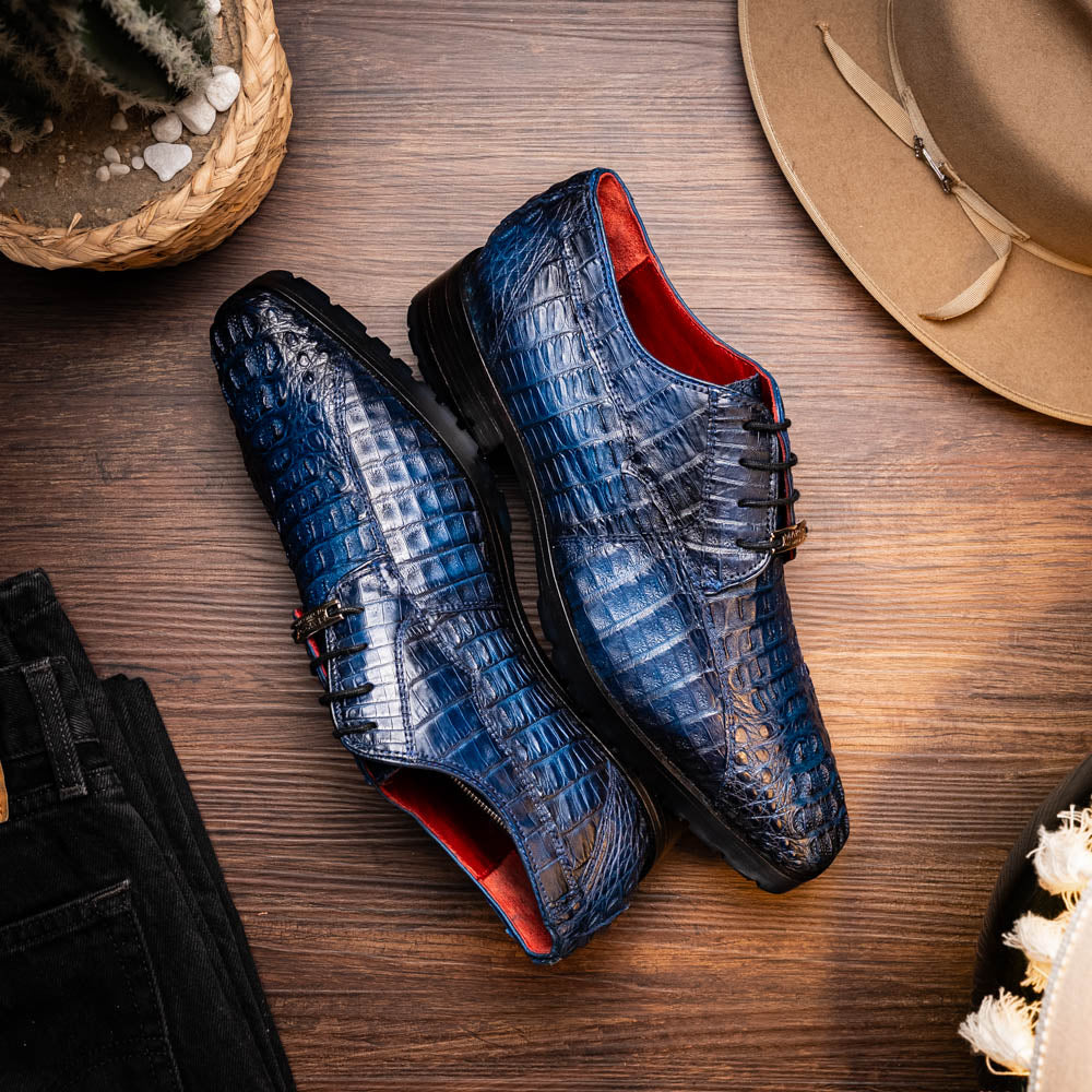 The Marco Di Milano Ragusa Stained Navy is a pair of luxury men's dress shoes in blue crocodile-patterned leather—one upright, one angled to display the sole—handcrafted for sophistication and timeless style.