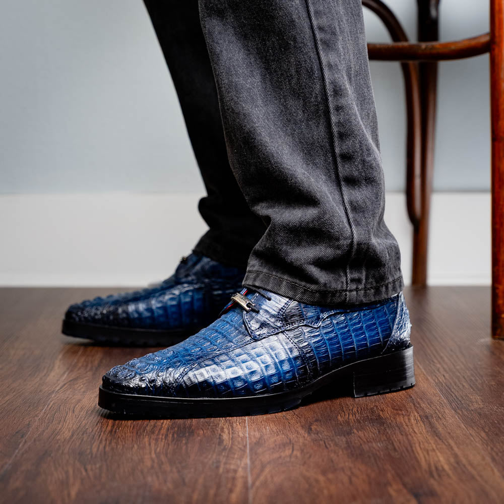 The Marco Di Milano Ragusa Stained Navy is a pair of luxury men's dress shoes in blue crocodile-patterned leather—one upright, one angled to display the sole—handcrafted for sophistication and timeless style.