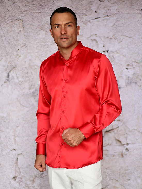 A man stands against a textured light-colored wall, wearing the Manzini MANZINI INR-102 Banded Collar Solid Color Satin Red Shirt and white pants, looking directly at the camera.