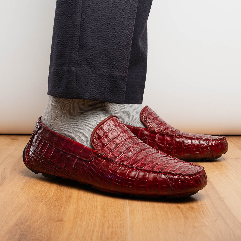 The Marco Di Milano Remo Wine features red crocodile-pattern leather driving shoes: one upright, one on its side showing a black rubber sole with red highlights—ideal for luxury men's footwear enthusiasts.
