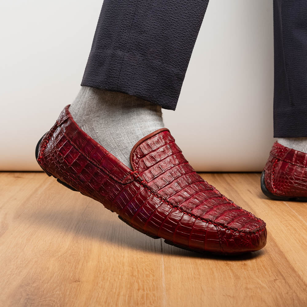 The Marco Di Milano Remo Wine features red crocodile-pattern leather driving shoes: one upright, one on its side showing a black rubber sole with red highlights—ideal for luxury men's footwear enthusiasts.