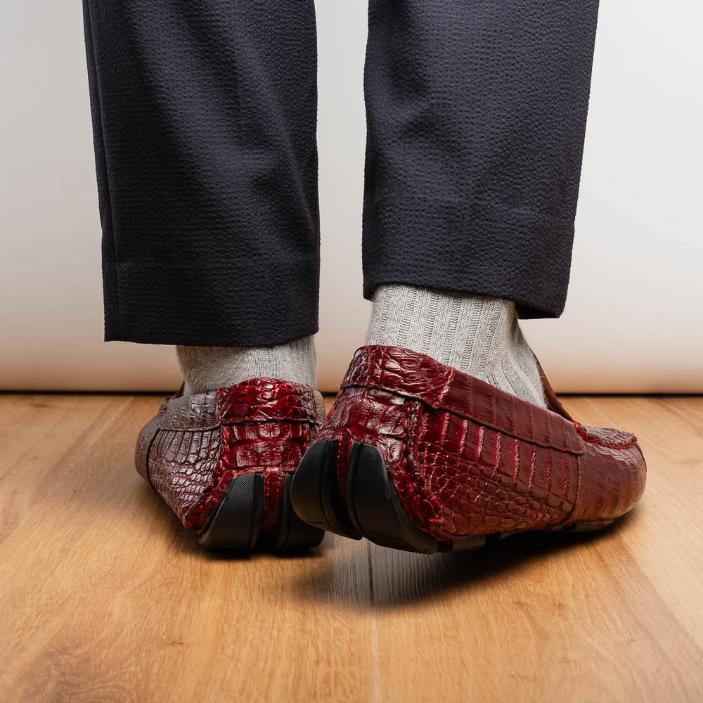 The Marco Di Milano Remo Wine features red crocodile-pattern leather driving shoes: one upright, one on its side showing a black rubber sole with red highlights—ideal for luxury men's footwear enthusiasts.