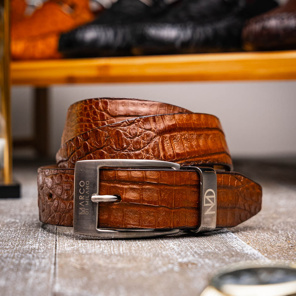 The Riccardo Belt Bay Apache by Marco Di Milano is a brown caiman belly belt with a crocodile pattern, featuring a silver buckle engraved with the brand name and "MM" initials—a true luxury exotic accessory.