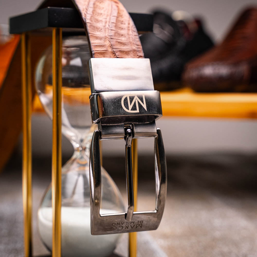 The Riccardo Belt Bay Apache by Marco Di Milano is a brown caiman belly belt with a crocodile pattern, featuring a silver buckle engraved with the brand name and "MM" initials—a true luxury exotic accessory.