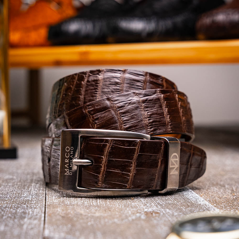 The Riccardo Belt Dark Brown by Marco Di Milano is crafted from exotic leather with a crocodile pattern and features a silver buckle engraved with the brand's name and logo.