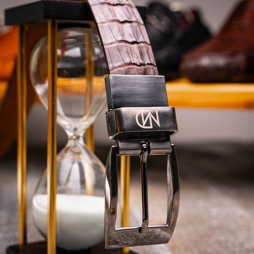 The Riccardo Belt Dark Brown by Marco Di Milano is crafted from exotic leather with a crocodile pattern and features a silver buckle engraved with the brand's name and logo.