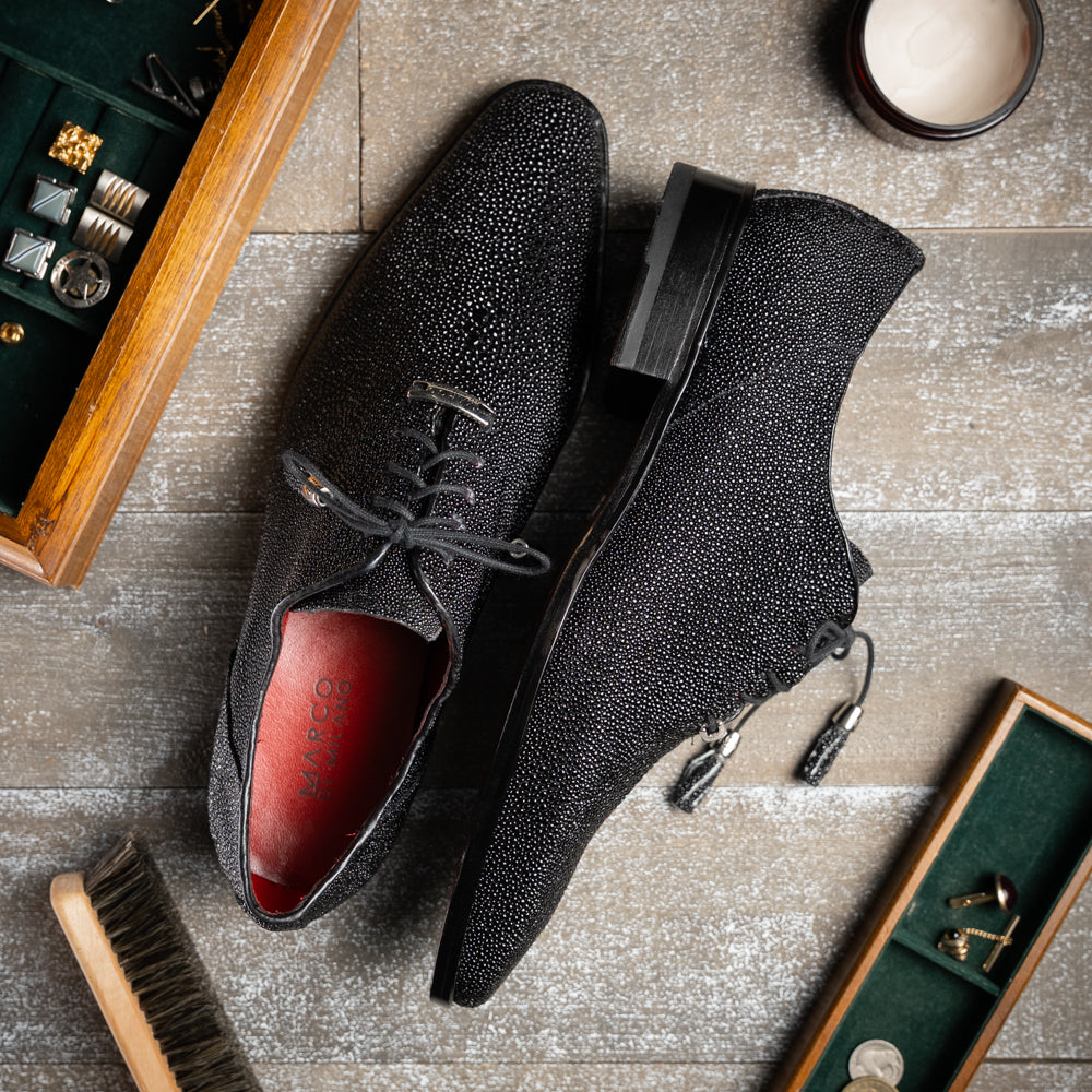 A pair of Rimini Black luxury dress shoes by Marco Di Milano, featuring a black textured finish and lace-up closure—one shoe displayed upright while the other shows the brown leather sole with embossed branding.