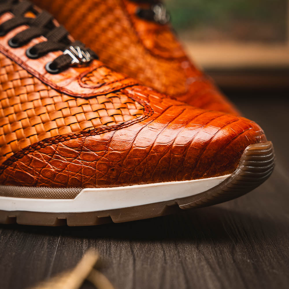 The Roma Brandy sneakers by Marco Di Milano feature brown woven calfskin, textured toe and heel, black laces, white midsoles, and brown rubber outsoles. One shoe stands upright while the other lies on its side.