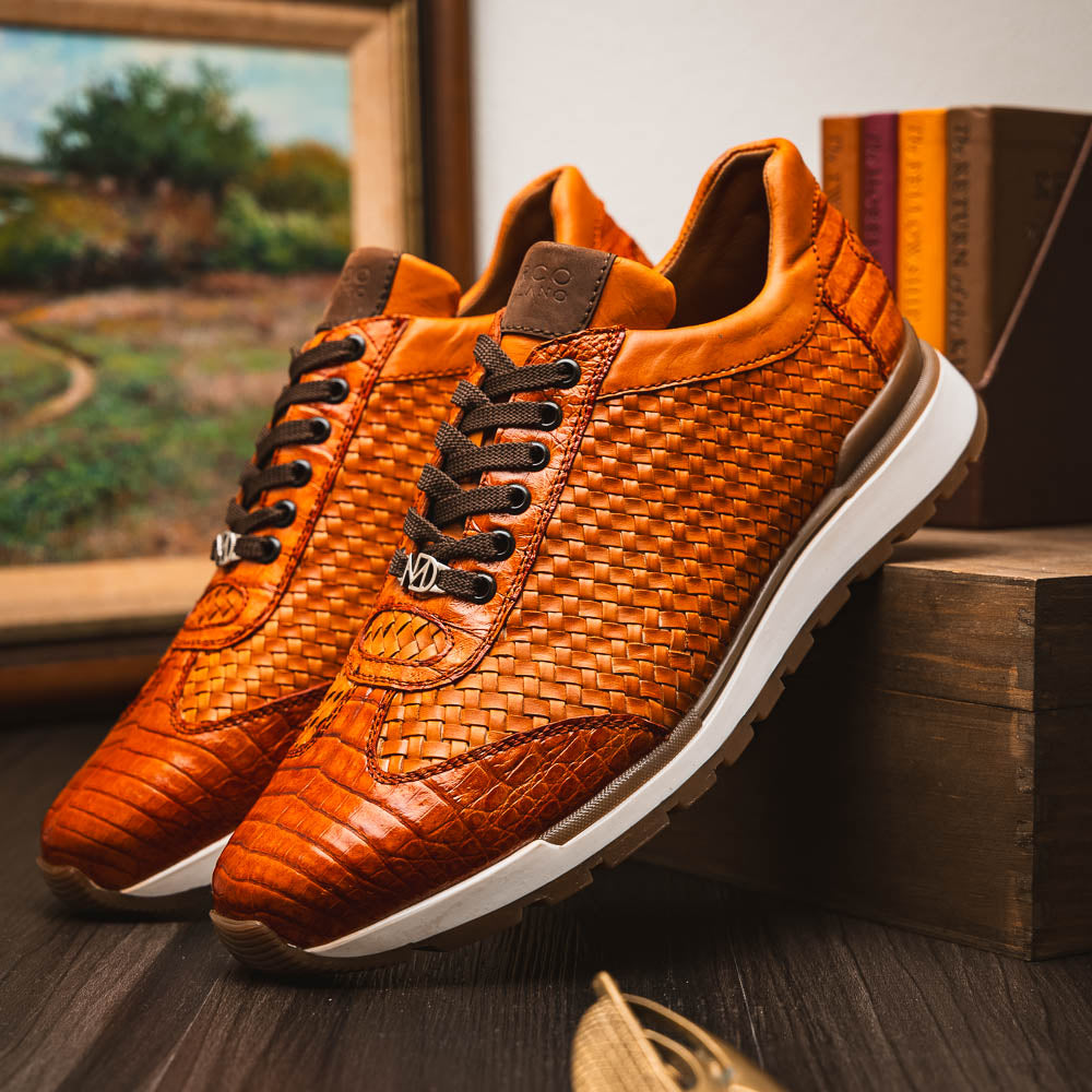 The Roma Brandy sneakers by Marco Di Milano feature brown woven calfskin, textured toe and heel, black laces, white midsoles, and brown rubber outsoles. One shoe stands upright while the other lies on its side.