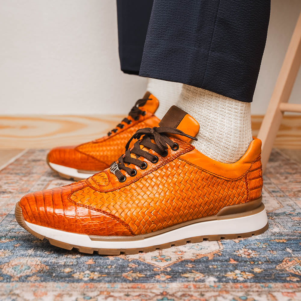 The Roma Brandy sneakers by Marco Di Milano feature brown woven calfskin, textured toe and heel, black laces, white midsoles, and brown rubber outsoles. One shoe stands upright while the other lies on its side.