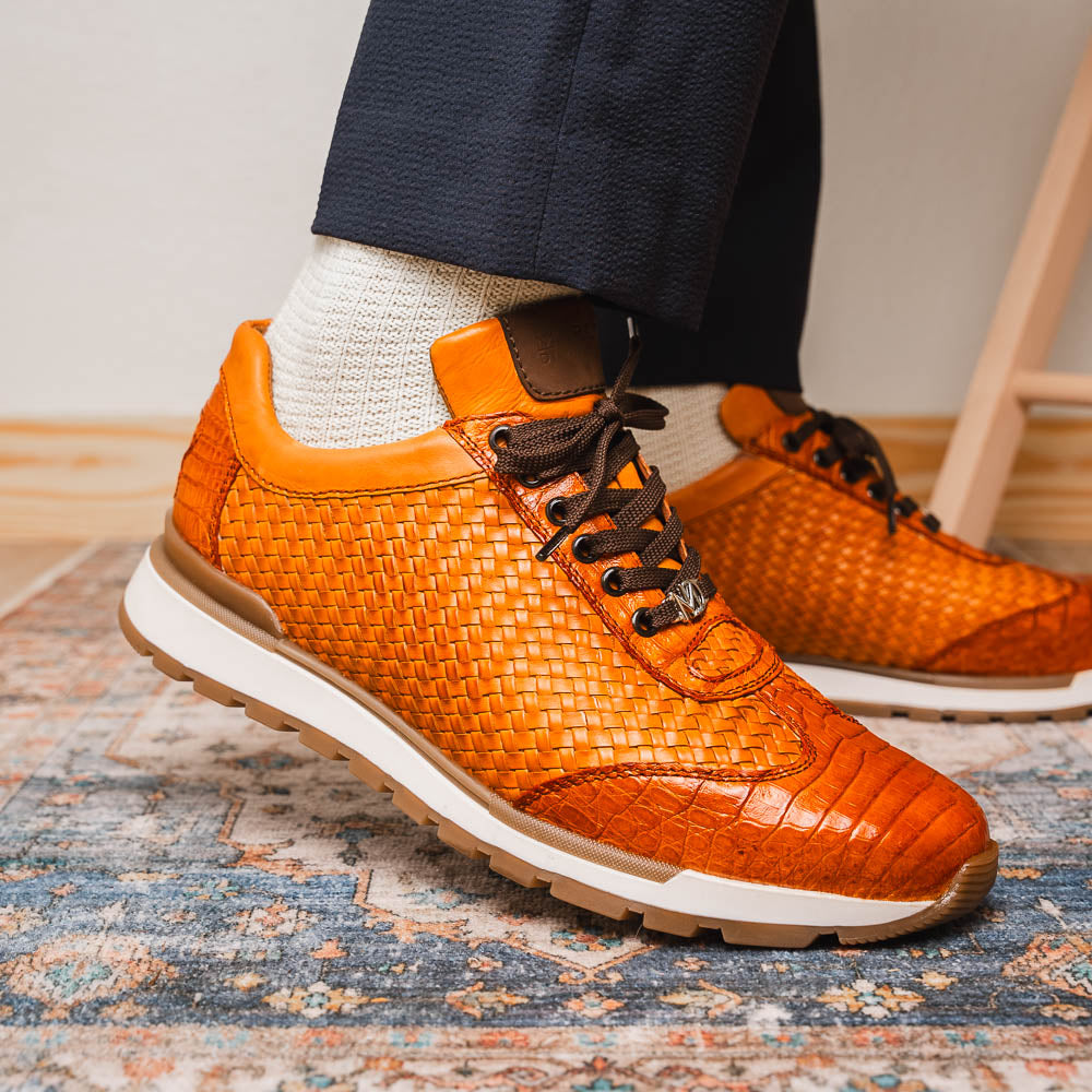 The Roma Brandy sneakers by Marco Di Milano feature brown woven calfskin, textured toe and heel, black laces, white midsoles, and brown rubber outsoles. One shoe stands upright while the other lies on its side.