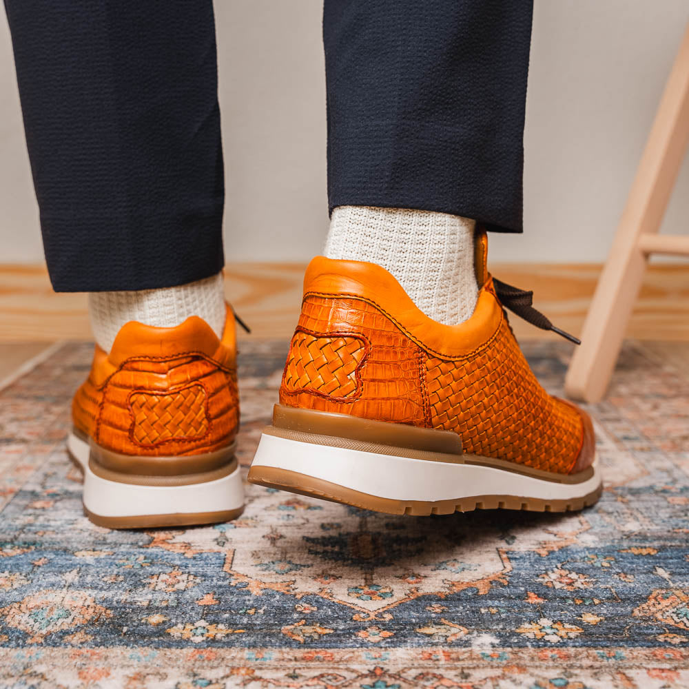 The Roma Brandy sneakers by Marco Di Milano feature brown woven calfskin, textured toe and heel, black laces, white midsoles, and brown rubber outsoles. One shoe stands upright while the other lies on its side.