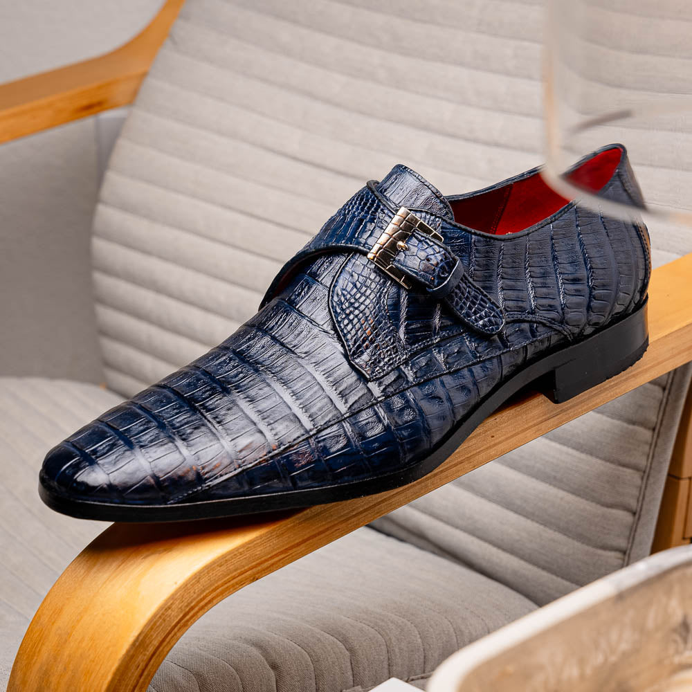 The Marco Di Milano Rovigo B Navy is a luxury men's monk strap shoe in blue crocodile-pattern leather, with one shoe upright and the other showing a tan sole stamped with the brand logo—perfect for dress shoe aficionados.