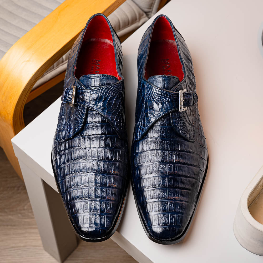 The Marco Di Milano Rovigo B Navy is a luxury men's monk strap shoe in blue crocodile-pattern leather, with one shoe upright and the other showing a tan sole stamped with the brand logo—perfect for dress shoe aficionados.