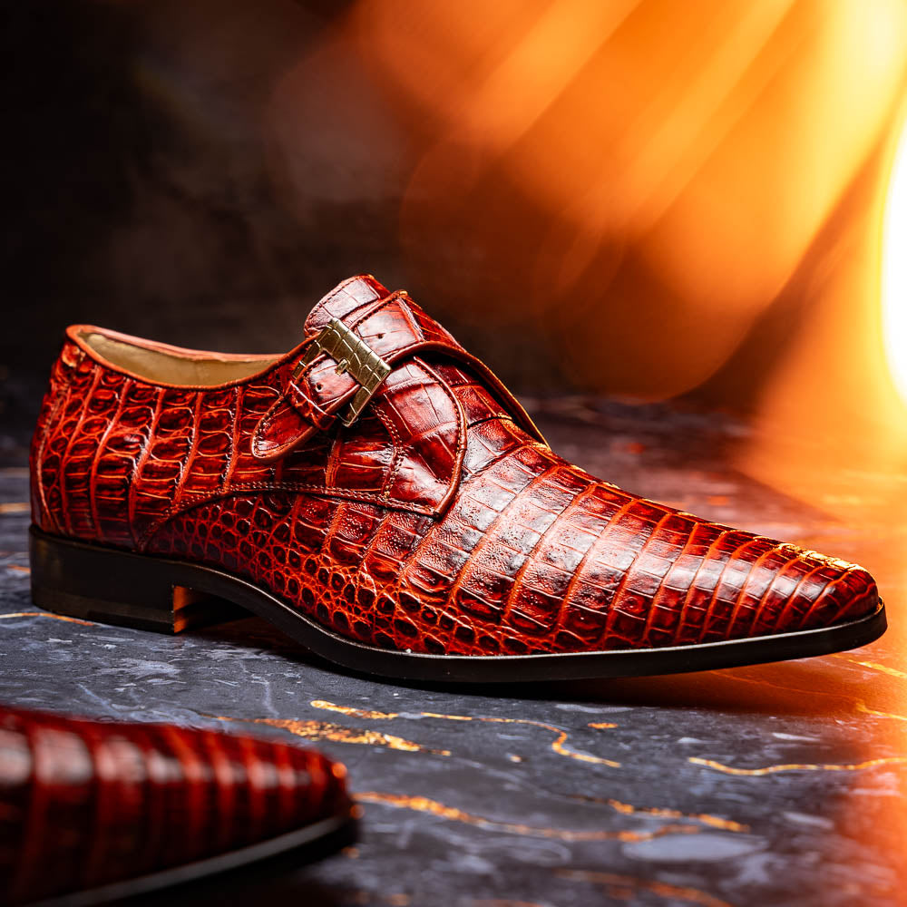 A pair of Rovigo Rustic Cognac monk strap dress shoes by Marco Di Milano, featuring shiny genuine caiman crocodile leather with a side buckle; one shoe is upright, and the other lies with its sole facing up.