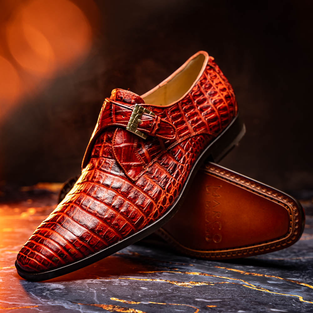 A pair of Rovigo Rustic Cognac monk strap dress shoes by Marco Di Milano, featuring shiny genuine caiman crocodile leather with a side buckle; one shoe is upright, and the other lies with its sole facing up.
