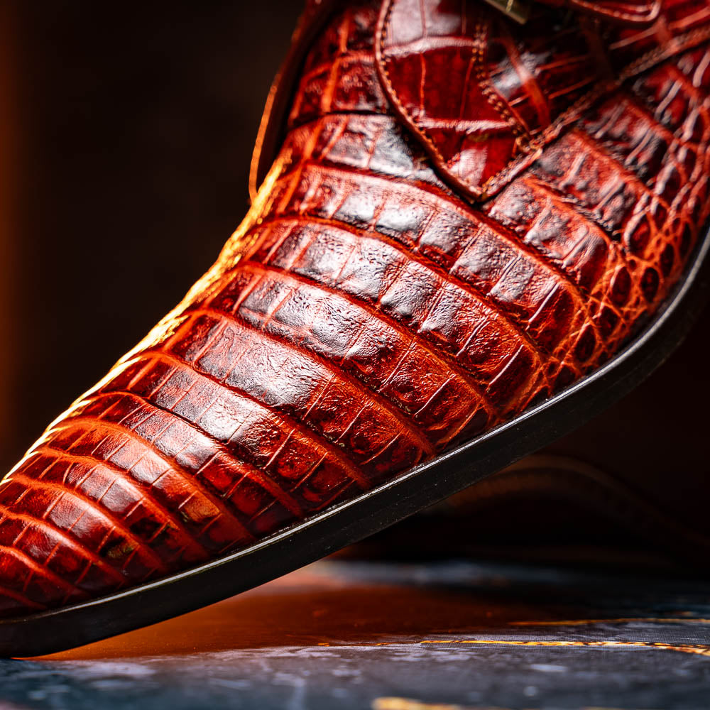 A pair of Rovigo Rustic Cognac monk strap dress shoes by Marco Di Milano, featuring shiny genuine caiman crocodile leather with a side buckle; one shoe is upright, and the other lies with its sole facing up.
