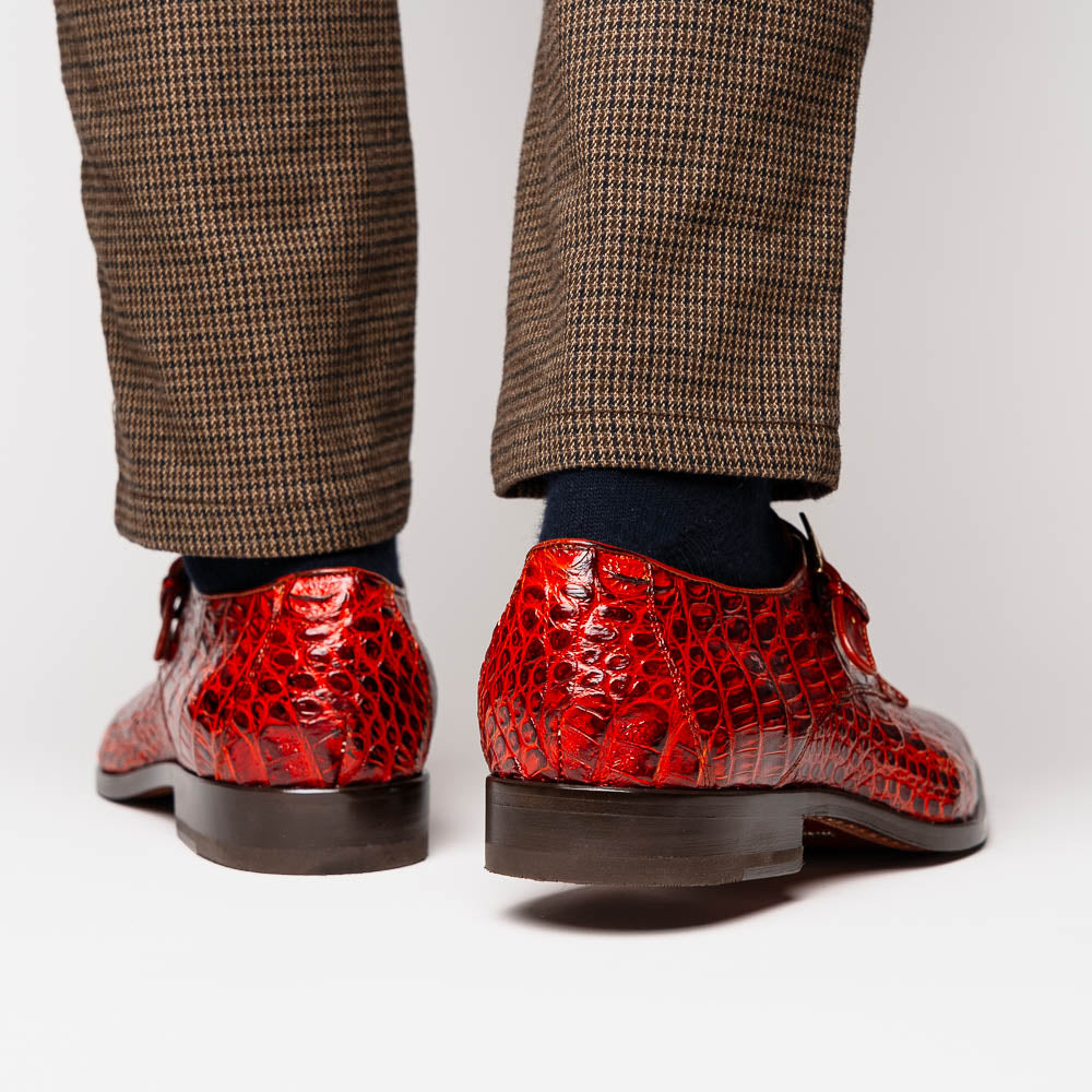 A pair of Rovigo Rustic Cognac monk strap dress shoes by Marco Di Milano, featuring shiny genuine caiman crocodile leather with a side buckle; one shoe is upright, and the other lies with its sole facing up.