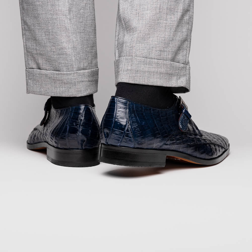 The Marco Di Milano Rovigo B Navy is a luxury men's monk strap shoe in blue crocodile-pattern leather, with one shoe upright and the other showing a tan sole stamped with the brand logo—perfect for dress shoe aficionados.