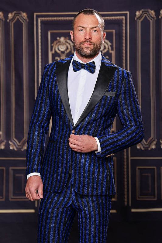 A man wears the MANZINI HEATHER Men's Slim-Fit 2-Piece Glitter Stripe Stage Prom Suit Royal by Manzini, featuring blue and black stripes, a black lapel, white shirt, and bow tie, standing before an ornate black and gold backdrop.