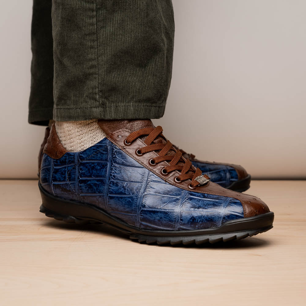 The Marco Di Milano Saulo Blue Jeans is a luxury men's sneaker featuring blue and brown hues, a textured crocodile-like pattern, and a black rubber sole, shown with one shoe upright and the other on its side.