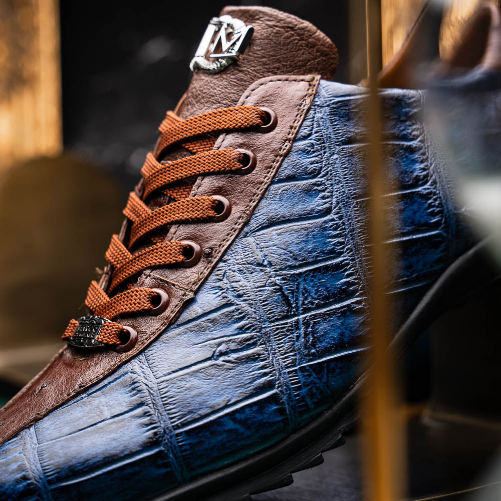 The Marco Di Milano Saulo Blue Jeans is a luxury men's sneaker featuring blue and brown hues, a textured crocodile-like pattern, and a black rubber sole, shown with one shoe upright and the other on its side.