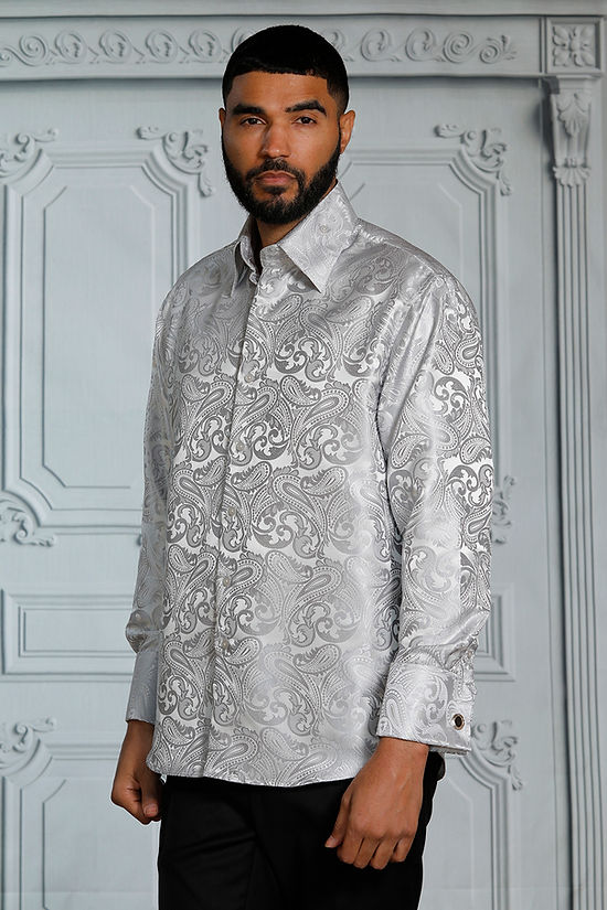 A man wears the Manzini MANZINI MZT-100 Paisley Long Sleeve Satin Shirt w/ Links in Silver, paired with black pants, standing before an ornate light gray wall—an elegant choice for formal occasions.