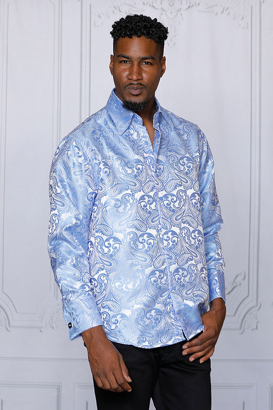 A man wears the Manzini MANZINI MZT-100 Paisley Long Sleeve Satin Shirt w/ Links in Sky and black pants, standing before a white ornate wall and looking at the camera with a neutral expression.