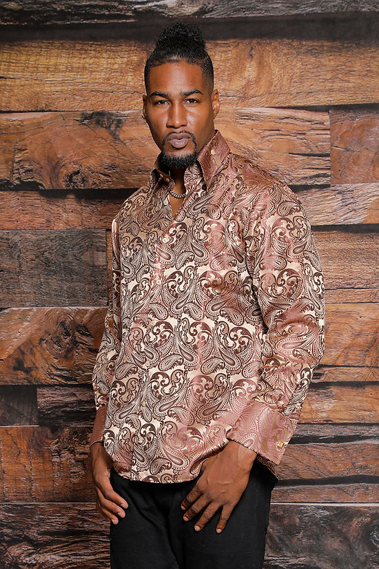 A man stands in front of a wooden plank wall, wearing the Manzini MANZINI MZT-100 Paisley Long Sleeve Satin Shirt with links in tan and black pants, facing forward with one hand in his pocket.