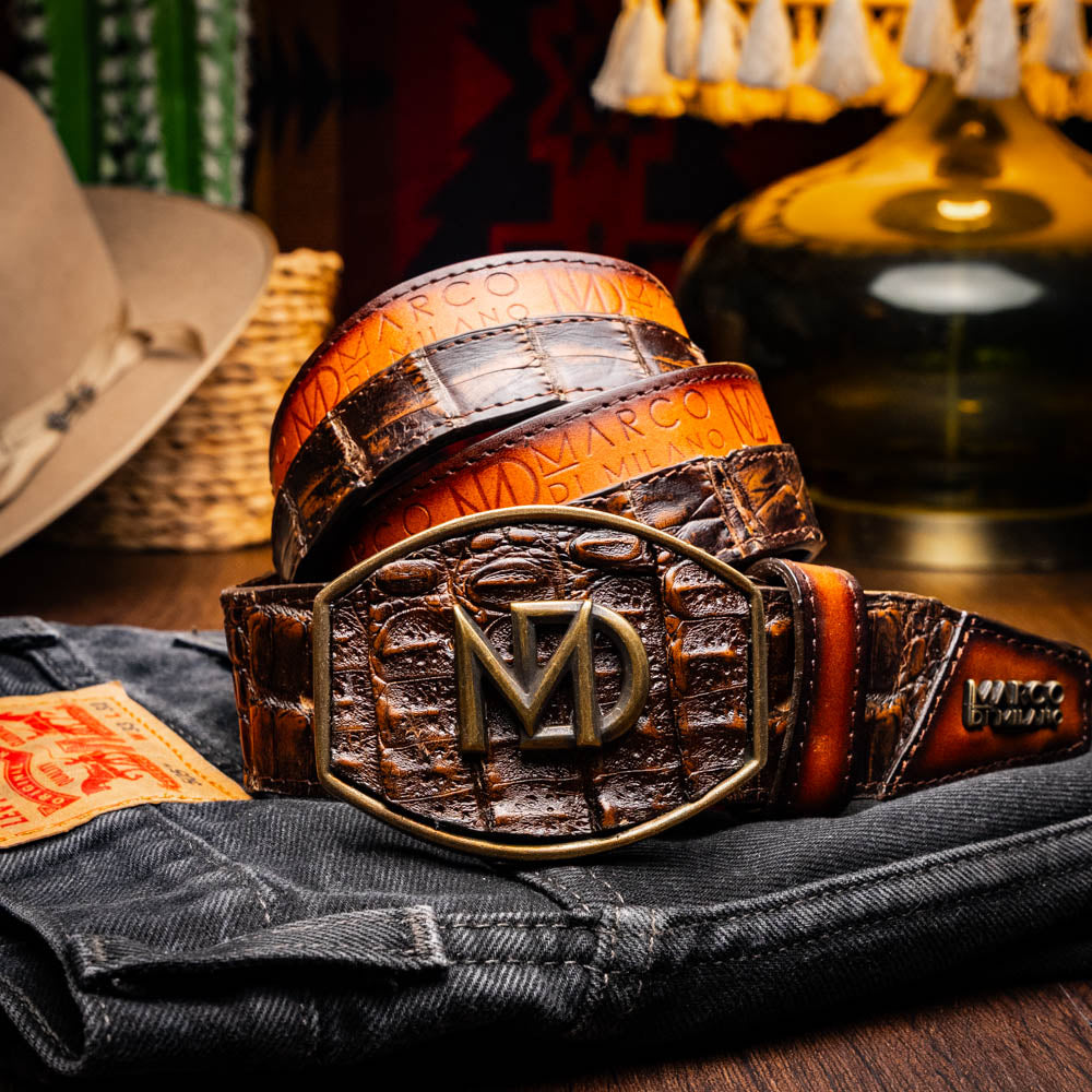 The Texas Belt Aged Brown by Marco Di Milano features embossed brown leather, a brass "MD" logo buckle, inner branding, and design details inspired by the elegance of a caiman crocodile belt.