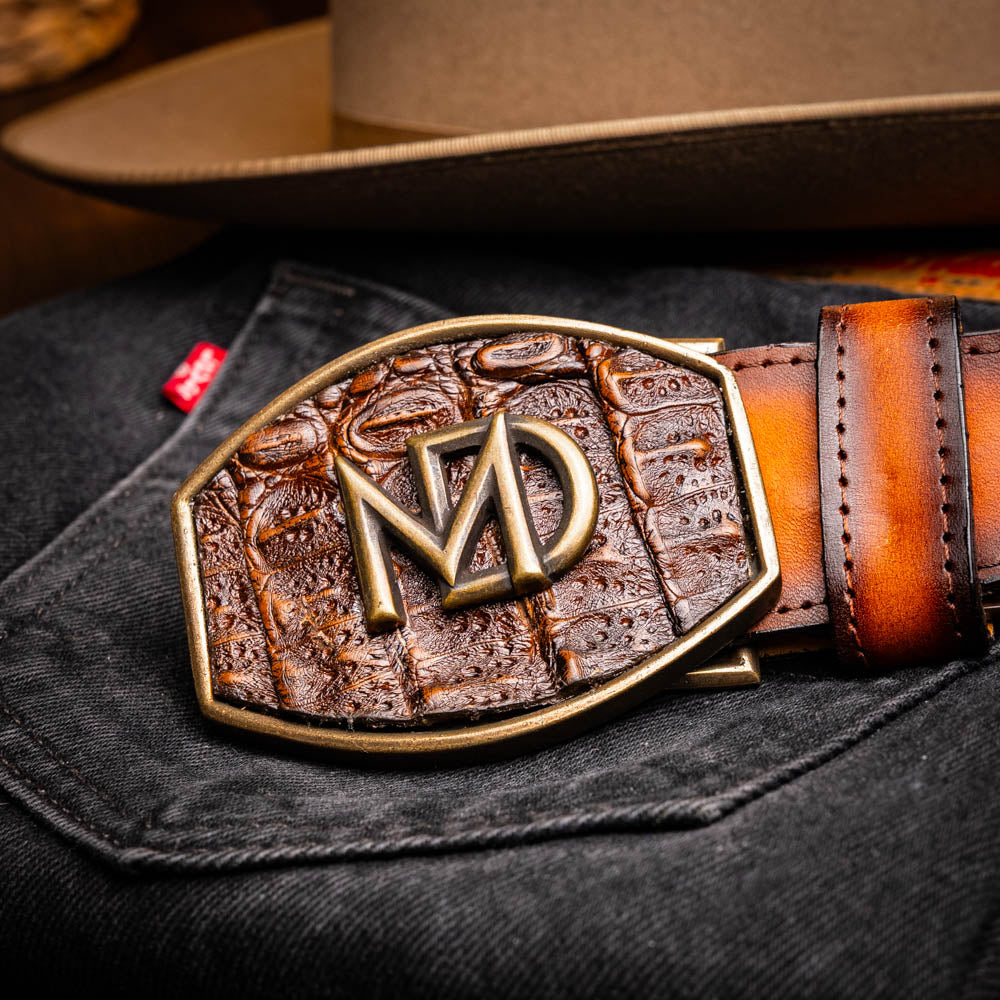 The Texas Belt Aged Brown by Marco Di Milano features embossed brown leather, a brass "MD" logo buckle, inner branding, and design details inspired by the elegance of a caiman crocodile belt.