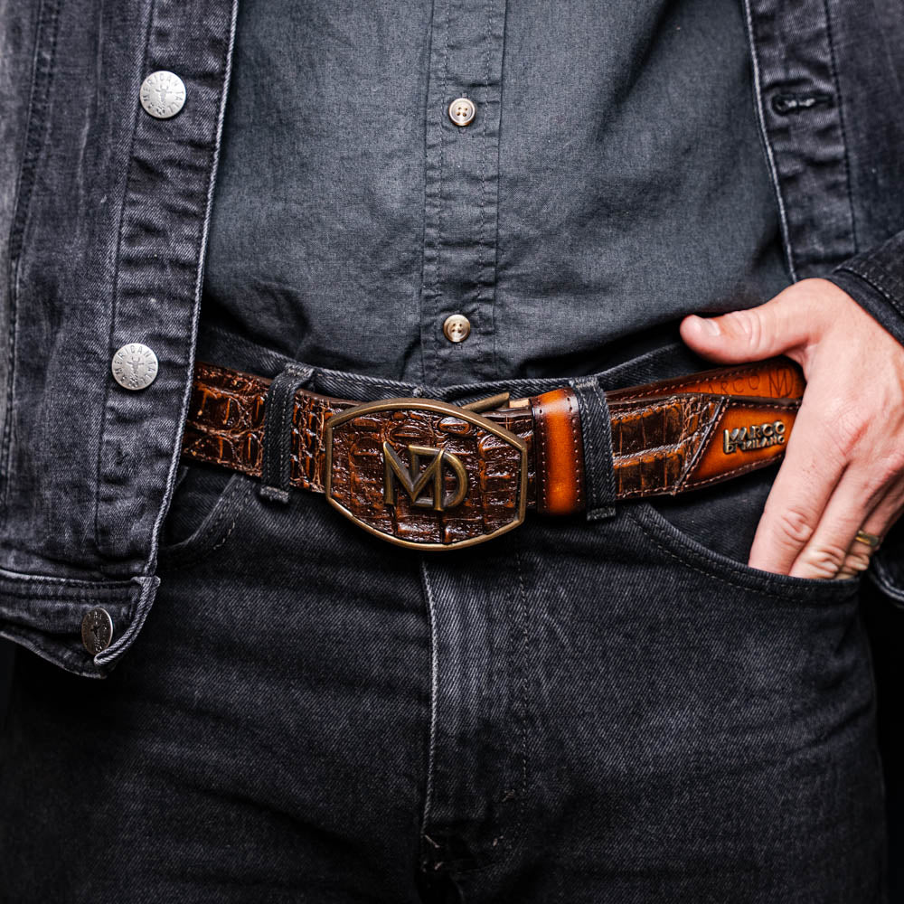 The Texas Belt Aged Brown by Marco Di Milano features embossed brown leather, a brass "MD" logo buckle, inner branding, and design details inspired by the elegance of a caiman crocodile belt.