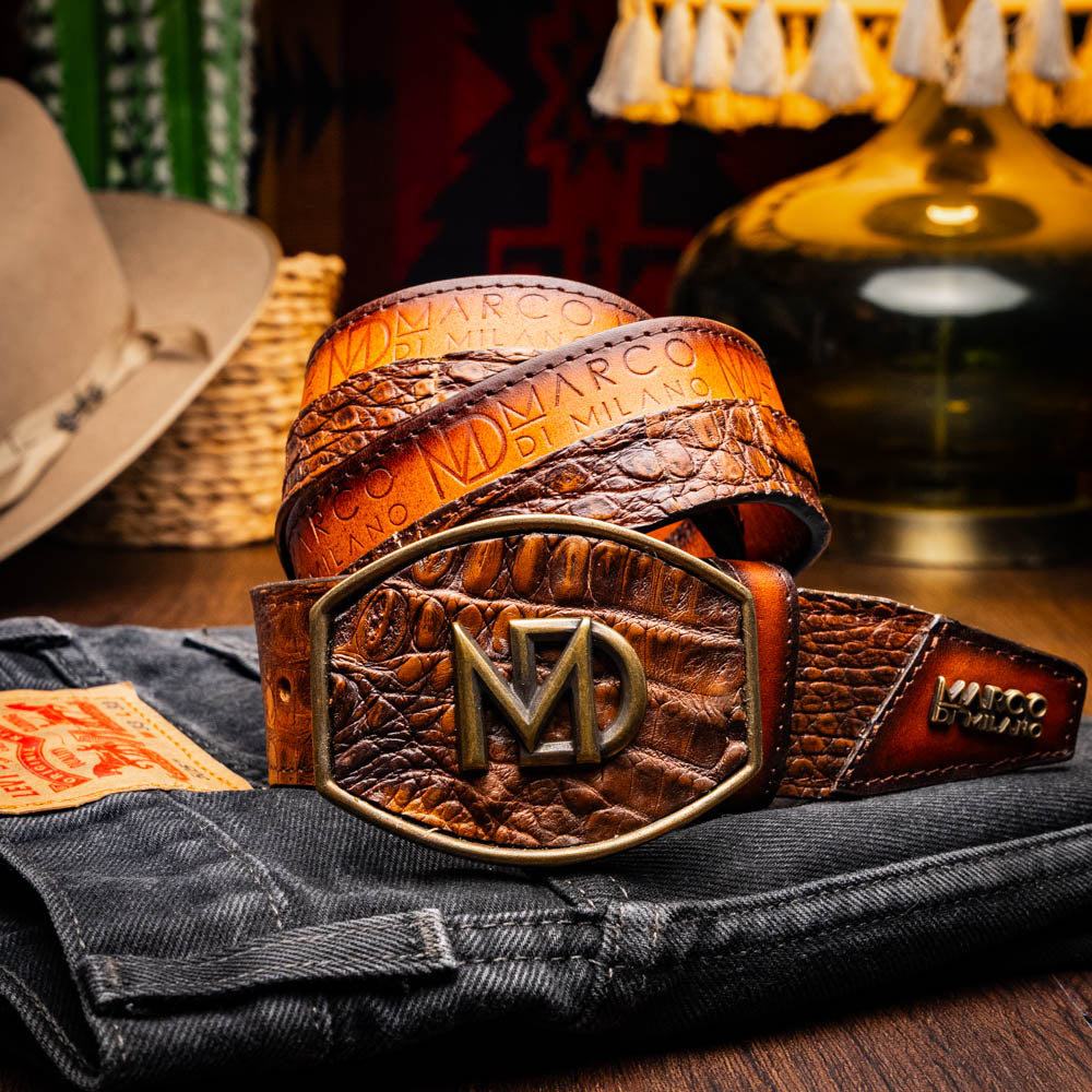 The Texas Belt Aged Maple by Marco Di Milano is crafted from luxurious caiman crocodile skin in a brown finish, featuring a large brass buckle engraved with "MD" and the brand name embossed on the leather.