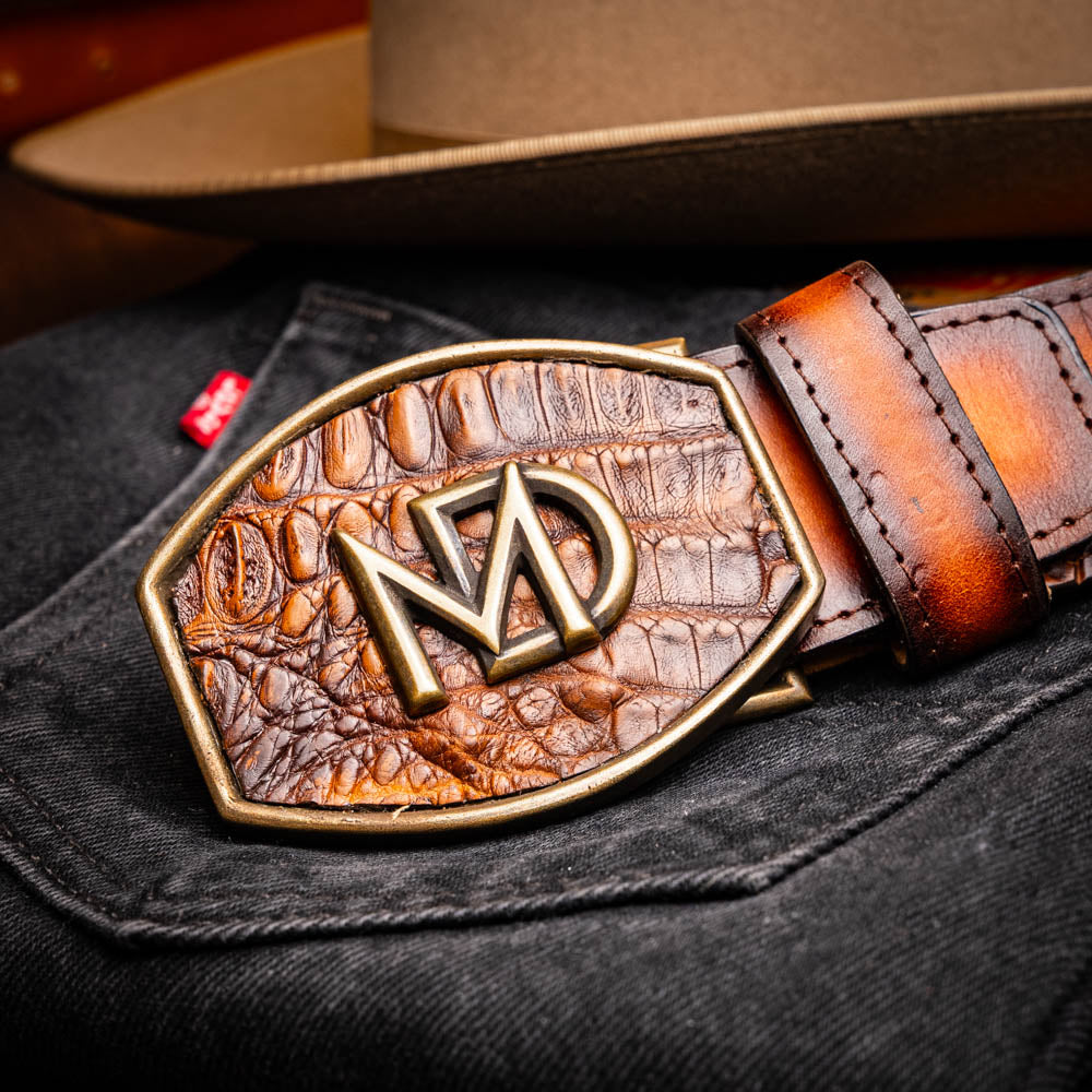 The Texas Belt Aged Maple by Marco Di Milano is crafted from luxurious caiman crocodile skin in a brown finish, featuring a large brass buckle engraved with "MD" and the brand name embossed on the leather.
