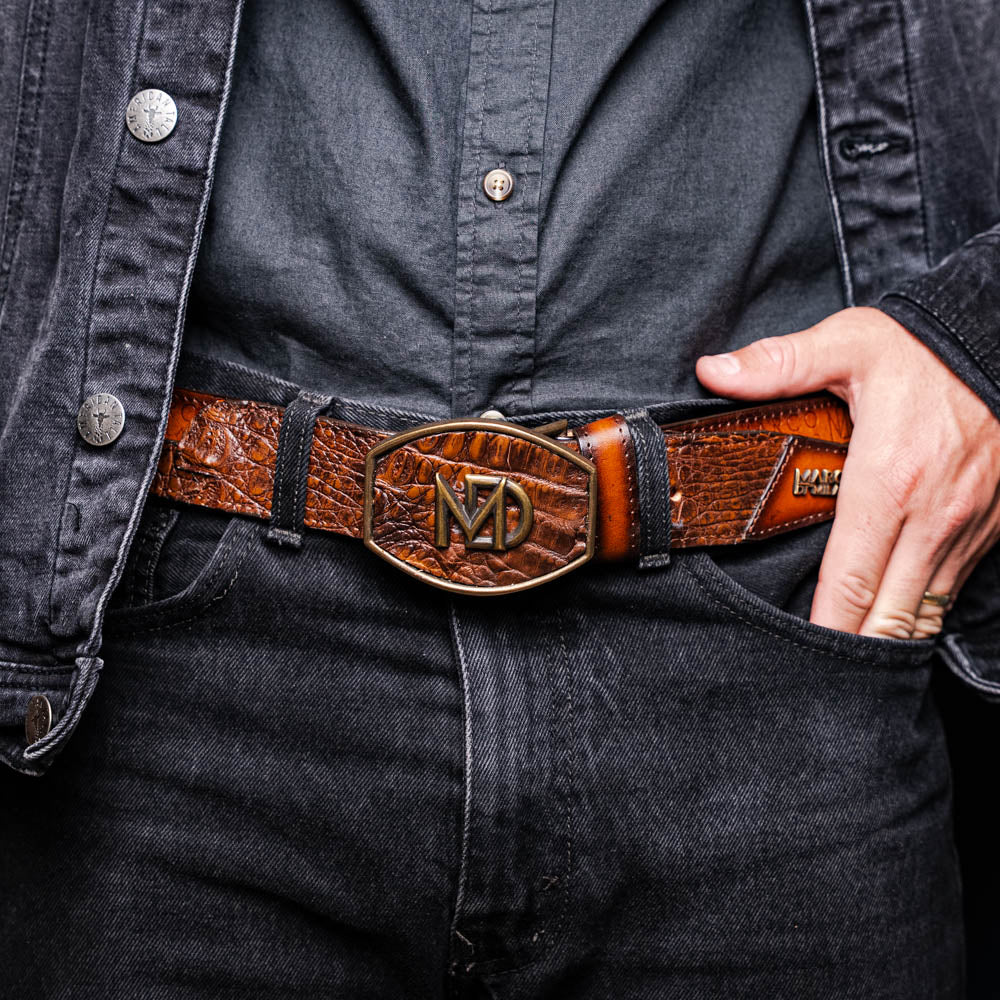 The Texas Belt Aged Maple by Marco Di Milano is crafted from luxurious caiman crocodile skin in a brown finish, featuring a large brass buckle engraved with "MD" and the brand name embossed on the leather.