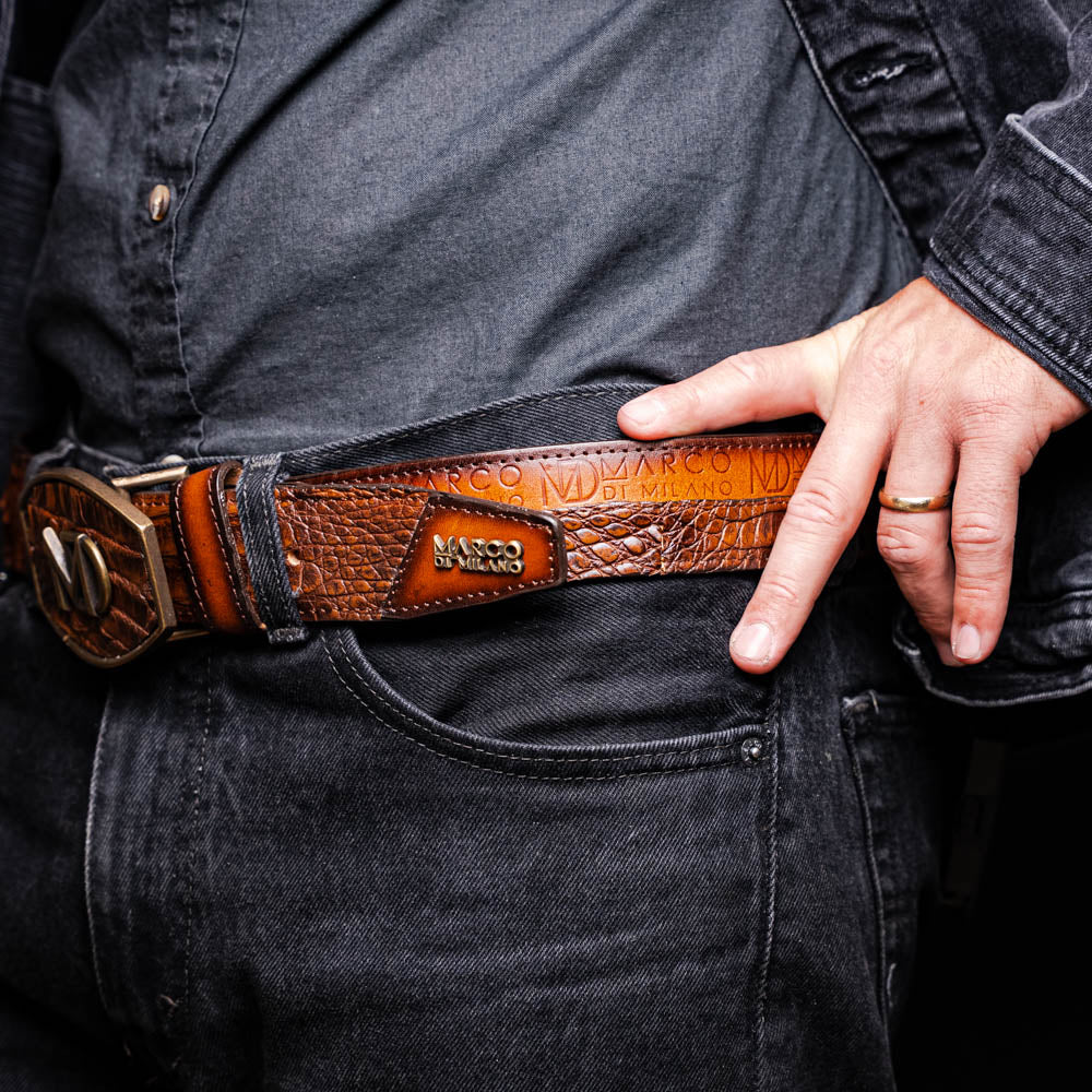 The Texas Belt Aged Maple by Marco Di Milano is crafted from luxurious caiman crocodile skin in a brown finish, featuring a large brass buckle engraved with "MD" and the brand name embossed on the leather.