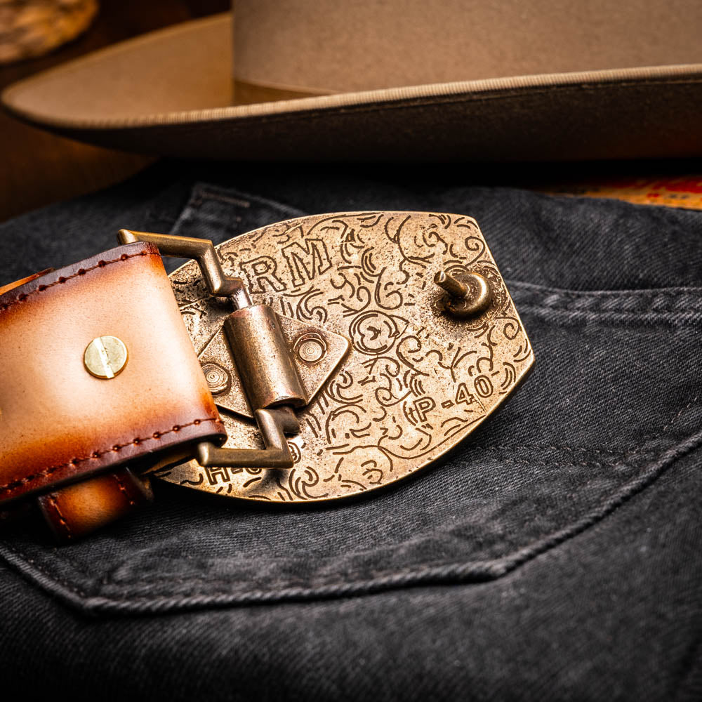 The Texas Belt Stone Brown by Marco Di Milano features embossed brown leather and a large gold-and-brown "CW" buckle, styled after a classic western calfskin belt with "MARCO DI MILANO" branding on the strap.